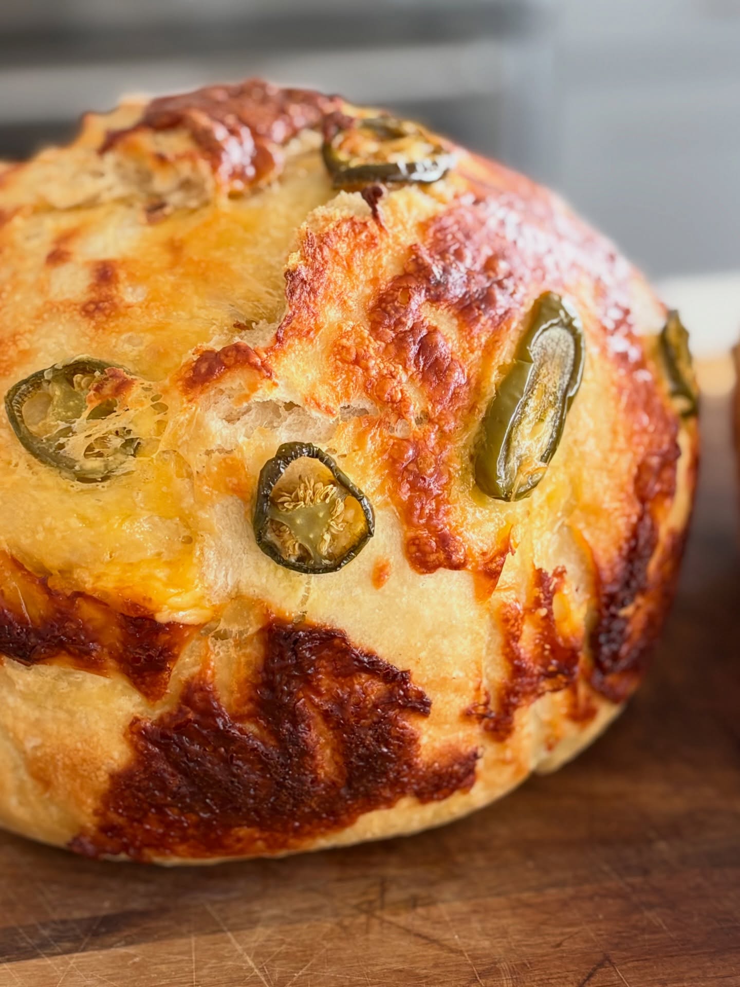 A fresh-baked golden jalapeño-cheddar artisan loaf, crust blistered and topped with jalapeño slices, on a wooden board